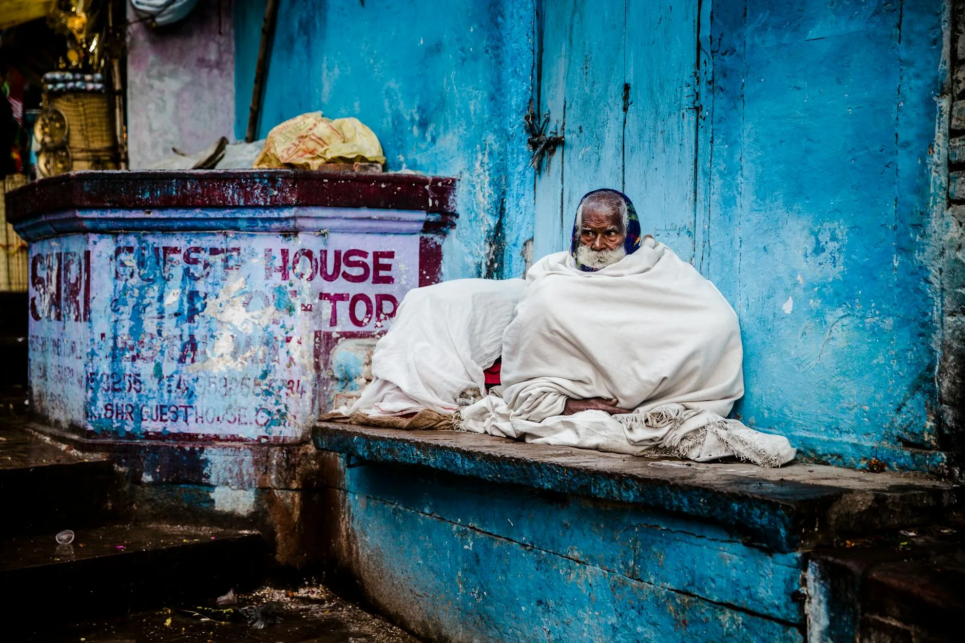 Starszy mężczyzna w białym kocu siedzi na ławce przy niebieskiej ścianie