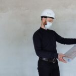 Architekt w kasku i maseczce przegląda plany budowlane w pomieszczeniu