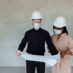Dwóch architektów w kaskach i maseczkach przegląda plany budowlane